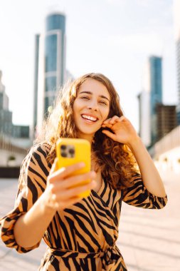 Güneşli bir caddede elinde telefonla duran gülümseyen bir kadının portresi. Güzel bir kadın blogu, metropol manzaralı bir gezintiden keyif alıyor. Blogculuk ve turizm kavramları.
