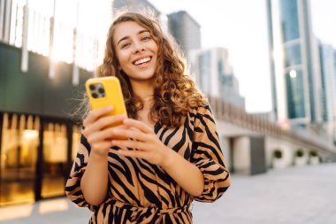 Güneşli bir caddede elinde telefonla duran gülümseyen bir kadının portresi. Güzel bir kadın blogu, metropol manzaralı bir gezintiden keyif alıyor. Blogculuk ve turizm kavramları.