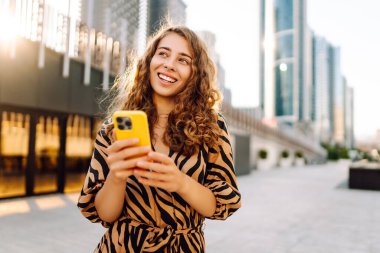 Güneşli bir caddede elinde telefonla duran gülümseyen bir kadının portresi. Güzel bir kadın blogu, metropol manzaralı bir gezintiden keyif alıyor. Blogculuk ve turizm kavramları.