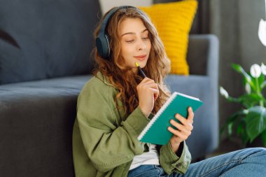 Gülümseyen bir kadın oturma odasında kablosuz kulaklıklar ve kitapla yerde oturuyor. Güzel bir kadın kitap okur ve samimi bir odada müzik dinler. Eğitim ve rahatlama kavramları.