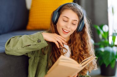 Gülümseyen bir kadın oturma odasında kablosuz kulaklıklar ve kitapla yerde oturuyor. Güzel bir kadın kitap okur ve samimi bir odada müzik dinler. Eğitim ve rahatlama kavramları.
