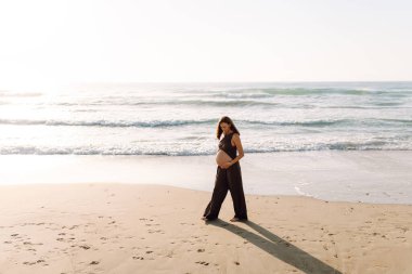 Çocuk bekleyen güzel bir kadın güneşin altında sahilde duruyor. Hamile kadın deniz manzarasını sever, gün batımını hisseder. Bekleme, yürüme, özgürlük ve rahatlama kavramları.