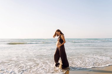 Çocuk bekleyen güzel bir kadın güneşin altında sahilde duruyor. Hamile kadın deniz manzarasını sever, gün batımını hisseder. Bekleme, yürüme, özgürlük ve rahatlama kavramları.