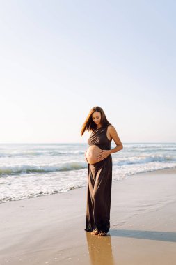 Beautiful woman expecting child stands on beach in sun's rays. Pregnant woman enjoys seascape, feeling freedom of sunset. Concepts of expecting, walking, freedom, and relaxation.