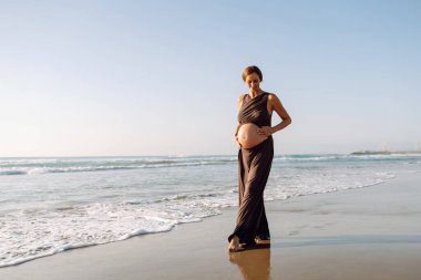 Çocuk bekleyen güzel bir kadın güneşin altında sahilde duruyor. Hamile kadın deniz manzarasını sever, gün batımını hisseder. Bekleme, yürüme, özgürlük ve rahatlama kavramları.