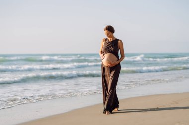 Çocuk bekleyen güzel bir kadın güneşin altında sahilde duruyor. Hamile kadın deniz manzarasını sever, gün batımını hisseder. Bekleme, yürüme, özgürlük ve rahatlama kavramları.