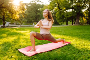 Neşeli bir kadın güneşli bir parkta minder üzerinde yoga yapıyor. Spor elbiseli bir kadın meditasyon yapar, dinlenir ya da zen 'i dışarıda bulur. Meditasyon, rahatlama kavramları.