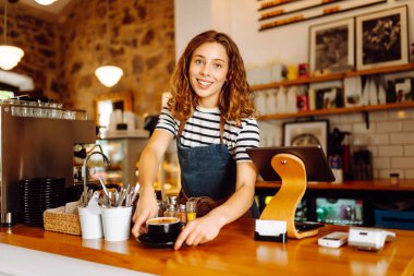 Kadın bir barista tezgahın arkasında duruyor, taze demlenmiş kahve servis ediyor. Önlüklü mutlu bir kadın sıcak bir kahve dükkanında aromatik kahve hazırlar. Küçük işletme konsepti, içkiler.