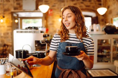 Kadın bir barista tezgahın arkasında duruyor, taze demlenmiş kahve servis ediyor. Önlüklü mutlu bir kadın sıcak bir kahve dükkanında aromatik kahve hazırlar. Küçük işletme konsepti, içkiler.