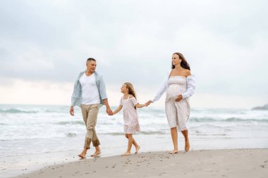 Bulutlu bir günde sahilde bebek bekleyen genç bir aile. Mutlu bir adam ve hamile bir kadın ve kızları birlikte vakit geçiriyorlar ve deniz manzarasına bakıyorlar. Aile ve sevgi kavramı.