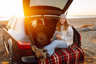 Bir kadın gezgin, şafak vakti bir arabanın bagajında otururken elinde termostan sıcak bir içecek tutar. Genç bir kadın yolculukta deniz manzarasından zevk alır. Macera kavramı, eğlence..