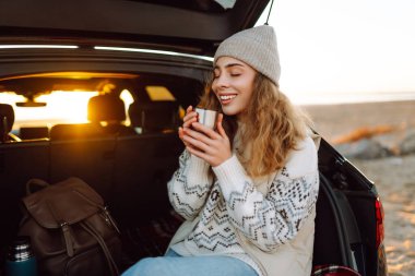 Bir kadın gezgin, şafak vakti bir arabanın bagajında otururken elinde termostan sıcak bir içecek tutar. Genç bir kadın yolculukta deniz manzarasından zevk alır. Macera kavramı, eğlence..
