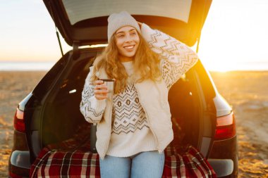 Bir kadın gezgin, şafak vakti bir arabanın bagajında otururken elinde termostan sıcak bir içecek tutar. Genç bir kadın yolculukta deniz manzarasından zevk alır. Macera kavramı, eğlence..