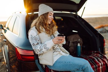 Bir kadın gezgin, şafak vakti bir arabanın bagajında otururken elinde termostan sıcak bir içecek tutar. Genç bir kadın yolculukta deniz manzarasından zevk alır. Macera kavramı, eğlence..