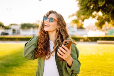 Güneşli bir parkta telefonu olan neşeli bir kadın. Güzel bir kadın temiz havada, bloglarda gezinir ve günbatımının tadını çıkarır. Bloglama kavramı, yürü.