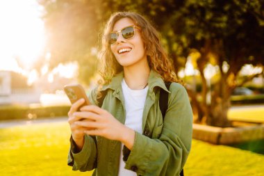 Güneşli bir parkta telefonu olan neşeli bir kadın. Güzel bir kadın temiz havada, bloglarda gezinir ve günbatımının tadını çıkarır. Bloglama kavramı, yürü.