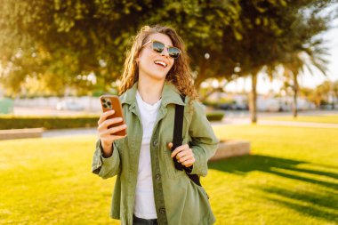 Güneşli bir parkta telefonu olan neşeli bir kadın. Güzel bir kadın temiz havada, bloglarda gezinir ve günbatımının tadını çıkarır. Bloglama kavramı, yürü.