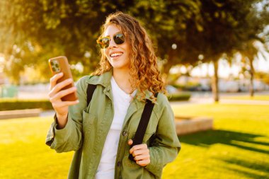 Güneşli bir parkta telefonu olan neşeli bir kadın. Güzel bir kadın temiz havada, bloglarda gezinir ve günbatımının tadını çıkarır. Bloglama kavramı, yürü.