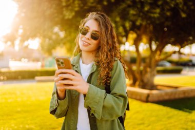 Güneşli bir parkta telefonu olan neşeli bir kadın. Güzel bir kadın temiz havada, bloglarda gezinir ve günbatımının tadını çıkarır. Bloglama kavramı, yürü.