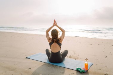 Spor elbiseli bir kadın plajda yoga minderinin üzerinde esniyor. Güzel bir kadın meditasyon yapar, dinlenir ya da deniz kenarında egzersiz yapar. Yoga, spor ve eğlence kavramları.