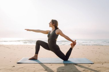 Spor elbiseli bir kadın plajda yoga minderinin üzerinde esniyor. Güzel bir kadın meditasyon yapar, dinlenir ya da deniz kenarında egzersiz yapar. Yoga, spor ve eğlence kavramları.