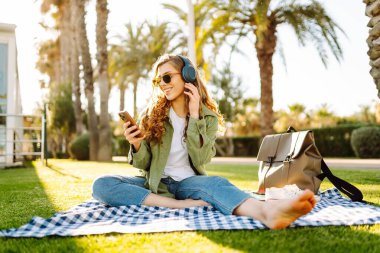 Genç bir kadın güneşli bir parkta telefon ve kablosuz kulaklıklarla oturuyor. Palmiye ağaçlarıyla çevrili. Güzel bir kadın gün batımında park çimlerinde dinlenmeyi sever. Müzik kavramı, rahatlama ve hafta sonları.