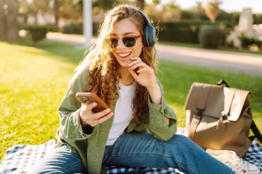 Genç bir kadın güneşli bir parkta telefon ve kablosuz kulaklıklarla oturuyor. Palmiye ağaçlarıyla çevrili. Güzel bir kadın gün batımında park çimlerinde dinlenmeyi sever. Müzik kavramı, rahatlama ve hafta sonları.