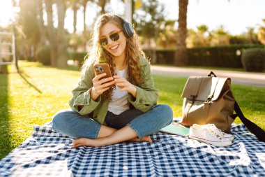 Genç bir kadın güneşli bir parkta telefon ve kablosuz kulaklıklarla oturuyor. Palmiye ağaçlarıyla çevrili. Güzel bir kadın gün batımında park çimlerinde dinlenmeyi sever. Müzik kavramı, rahatlama ve hafta sonları.