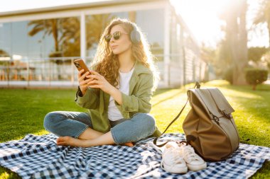 Genç bir kadın güneşli bir parkta telefon ve kablosuz kulaklıklarla oturuyor. Palmiye ağaçlarıyla çevrili. Güzel bir kadın gün batımında park çimlerinde dinlenmeyi sever. Müzik kavramı, rahatlama ve hafta sonları.