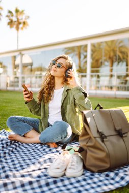 Genç bir kadın güneşli bir parkta telefon ve kablosuz kulaklıklarla oturuyor. Palmiye ağaçlarıyla çevrili. Güzel bir kadın gün batımında park çimlerinde dinlenmeyi sever. Müzik kavramı, rahatlama ve hafta sonları.