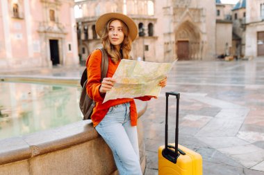 Şapkalı ve parlak bavullu güzel bir turist güneşli şehir meydanındaki fıskiyenin yanında elinde haritayla duruyor. Sırt çantalı kadın yeni şehri keşfetmekten zevk alıyor. Turizm, navigasyon, tatil kavramları.