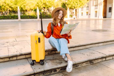 Şapkalı ve parlak bavullu güzel bir turist güneşli şehir meydanındaki fıskiyenin yanında elinde haritayla duruyor. Sırt çantalı kadın yeni şehri keşfetmekten zevk alıyor. Turizm, navigasyon, tatil kavramları.