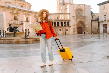 Şapkalı ve parlak bavullu güzel bir turist güneşli şehir meydanındaki fıskiyenin yanında elinde haritayla duruyor. Sırt çantalı kadın yeni şehri keşfetmekten zevk alıyor. Turizm, navigasyon, tatil kavramları.