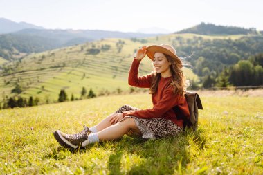 Şapka takan ve sırt çantası taşıyan genç bir kadın yeşil tepede güneşli bir günün tadını çıkarıyor. Gezgin, yürüyüş yolunda yürürken ve dağ manzarasına bakarken kendini özgür hissediyor. Kavram: macera, rahatlama.