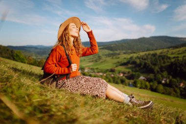 Şapka takan ve sırt çantası taşıyan genç bir kadın yeşil tepede güneşli bir günün tadını çıkarıyor. Gezgin, yürüyüş yolunda yürürken ve dağ manzarasına bakarken kendini özgür hissediyor. Kavram: macera, rahatlama.