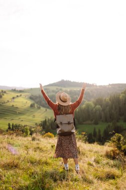 Şapka takan ve sırt çantası taşıyan genç bir kadın yeşil tepede güneşli bir günün tadını çıkarıyor. Gezgin, yürüyüş yolunda yürürken ve dağ manzarasına bakarken kendini özgür hissediyor. Kavram: macera, rahatlama.