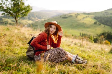 Şapka takan ve sırt çantası taşıyan genç bir kadın yeşil tepede güneşli bir günün tadını çıkarıyor. Gezgin, yürüyüş yolunda yürürken ve dağ manzarasına bakarken kendini özgür hissediyor. Kavram: macera, rahatlama.