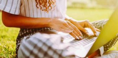 Genç bayan serbest çalışan dizüstü bilgisayarda çalışırken parktaki yeşil çayır üzerinde telefonla oturuyor. Kablosuz kulaklıklı mutlu kadın dışarıda dizüstü bilgisayar kullanıyor. Teknoloji ve rahatlama kavramı.