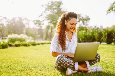 Genç bayan serbest çalışan dizüstü bilgisayarda çalışırken parktaki yeşil çayır üzerinde telefonla oturuyor. Kablosuz kulaklıklı mutlu kadın dışarıda dizüstü bilgisayar kullanıyor. Teknoloji ve rahatlama kavramı.