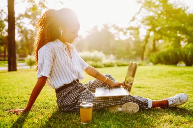 Genç bayan serbest çalışan dizüstü bilgisayarda çalışırken parktaki yeşil çayır üzerinde telefonla oturuyor. Kablosuz kulaklıklı mutlu kadın dışarıda dizüstü bilgisayar kullanıyor. Teknoloji ve rahatlama kavramı.