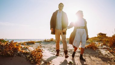 A stylish young couple strolls together along the beach at sunset. The young woman and man enjoy the freedom of the seascape in the sun's rays. Conceptual images of love, nature, and relaxation.