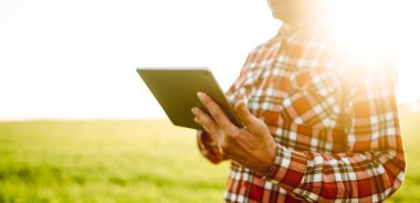 Gün batımında elinde dijital tablet olan yeşil bir tarla çiftçisinin yakın çekimi. Bir tarım uzmanı dijital tablet üzerinde çalışır ve ürün büyümesini kontrol eder. Çiftçilik ve teknoloji kavramları.