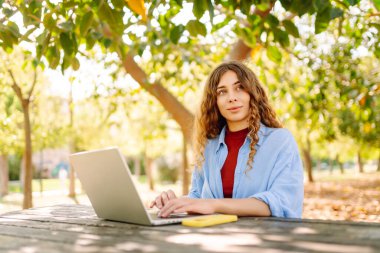Gülümseyen bir kadın parkta bir masada oturuyor ve dizüstü bilgisayarla çalışıyor. Dişi bir serbest yazar dışarıda dizüstü bilgisayarıyla güneşli havanın tadını çıkarır. Uzaktan çalışma, rahatlama ve hafta sonları kavramı.