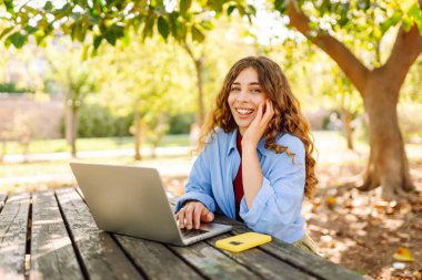 A smiling woman sits at a table in a park, working on a laptop. A female freelancer enjoys the sunny weather with her laptop outdoors. Concept of remote work, relaxation, and weekends.
