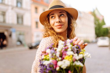 Şirin bir kadın bir buket kır çiçeğiyle güneşli bir şehir caddesinde geziniyor. Şapkalı güzel bir turist mimariyi, ellerinde çiçeklerle şehrin atmosferini seviyor. Kutlama, rahatlama kavramları