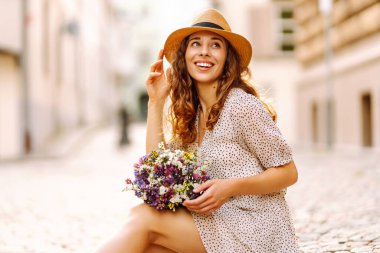 Şirin bir kadın bir buket kır çiçeğiyle güneşli bir şehir caddesinde geziniyor. Şapkalı güzel bir turist mimariyi, ellerinde çiçeklerle şehrin atmosferini seviyor. Kutlama, rahatlama kavramları