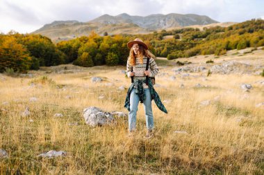 Sırt çantası ve şapkası olan dağ yürüyüşü yolunda genç bir yürüyüşçü. Gülümseyen bir kadın gün batımında dağ manzarasının keyfini çıkarıyor. Hafta sonları, özgürlük ve macera kavramları.