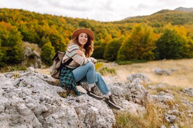 Sırt çantası ve şapkası olan dağ yürüyüşü yolunda genç bir yürüyüşçü. Gülümseyen bir kadın gün batımında dağ manzarasının keyfini çıkarıyor. Hafta sonları, özgürlük ve macera kavramları.