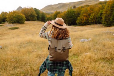 Sırt çantası ve şapkası olan dağ yürüyüşü yolunda genç bir yürüyüşçü. Gülümseyen bir kadın gün batımında dağ manzarasının keyfini çıkarıyor. Hafta sonları, özgürlük ve macera kavramları.