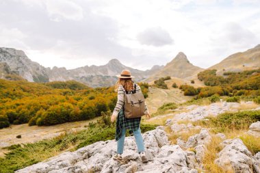 Sırt çantası ve şapkası olan dağ yürüyüşü yolunda genç bir yürüyüşçü. Gülümseyen bir kadın gün batımında dağ manzarasının keyfini çıkarıyor. Hafta sonları, özgürlük ve macera kavramları.